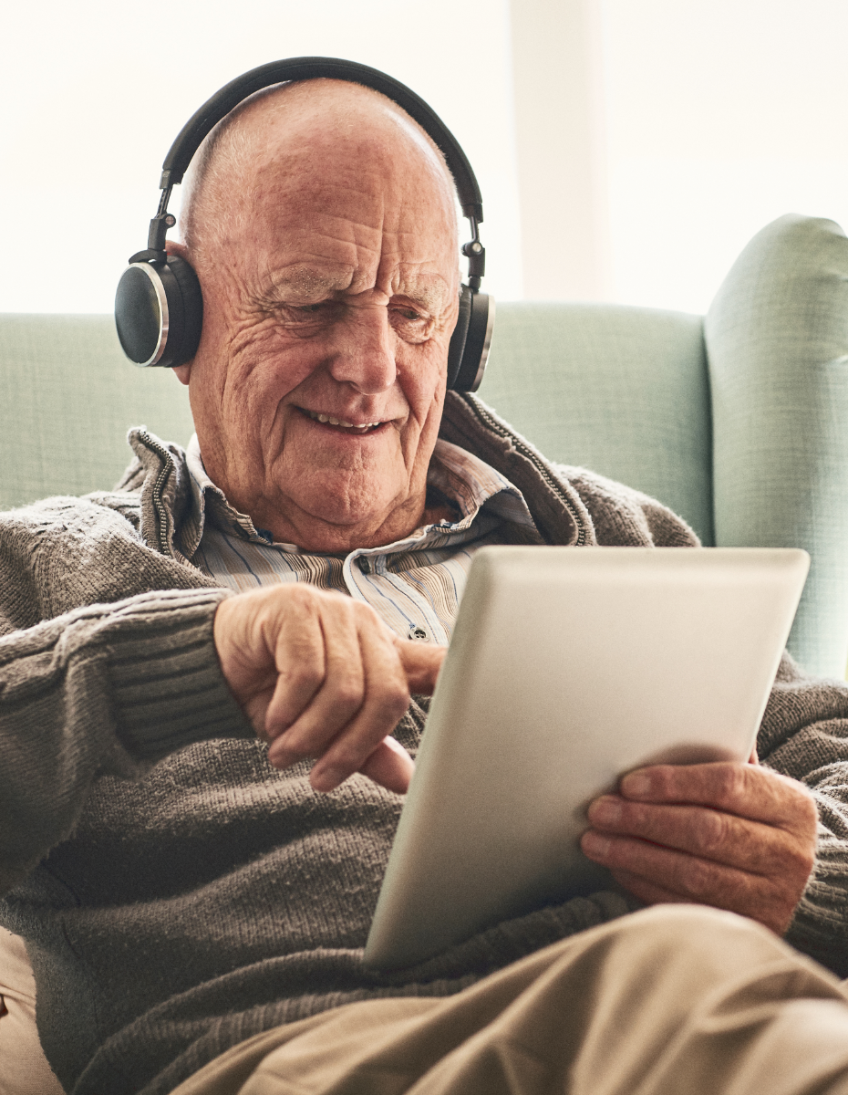 Älterer Mann sitzt mit Kopfhörern auf dem Sofa uns schaut in Tablet.